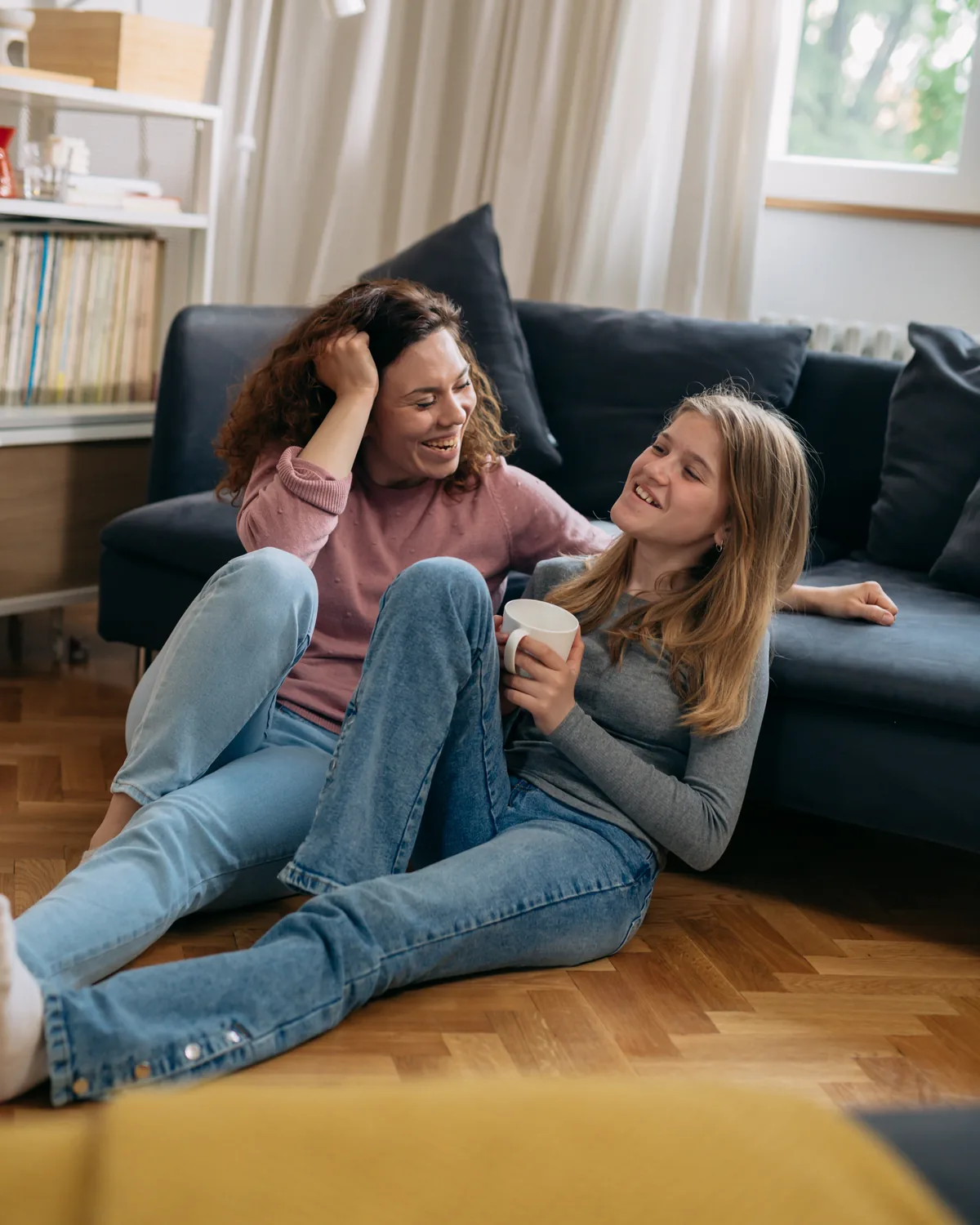 A mother and her teenage daughter sitting on the floor, talking and laughing.